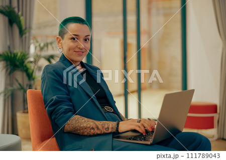 Smiling tattooed female freelancer with green short hair working on laptop while sitting in office Smiling tattooed female freelancer with green short hair working on laptop while sitting in office 111789043