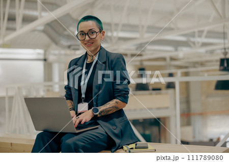 Pretty tattooed female manager is use laptop sitting in modern coworking and looks camera Pretty tattooed female manager is use laptop sitting in modern coworking and looks camera 111789080