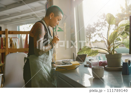 Woman in apron views order details in notebook during working day in design studio Woman in apron views order details in notebook during working day in design studio 111789140