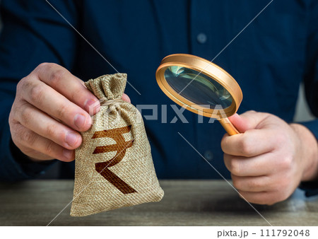 A man examines a indian rupee money bag through a magnifying glass. Investigating capital origins. Anti money laundering, tax evasion. Study terms and conditions on deposit or loan. 111792048