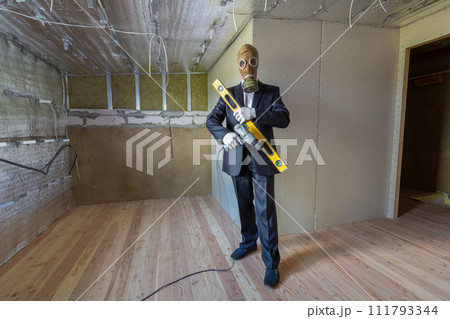 Strange man in businessman suit and gas protection mask inside a room under renovation works holding electric screwdriver and a level tools. 111793344