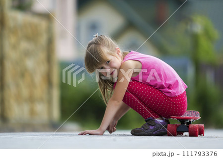 Pretty young long-haired blond child girl in casual pink clothing sits leaning on hands on skateboard on sunny street on blurred summer green background. Children activities, games and fun concept. Pretty young long-haired blond child girl in casual pink clothing sits leaning on hands on skateboard on sunny street on blurred summer green background. Children activities, games and fun concept. 111793376