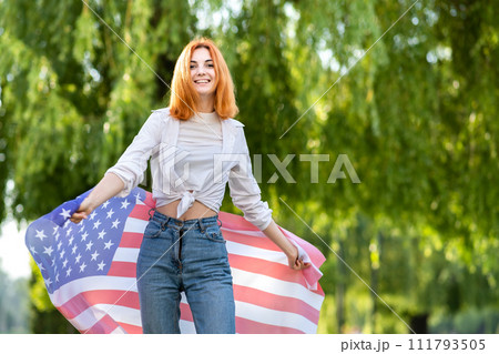 Happy young red haired woman posing with USA national flag standing outdoors in summer park. Positive girl celebrating United States independence day. Happy young red haired woman posing with USA national flag standing outdoors in summer park. Positive girl celebrating United States independence day. 111793505
