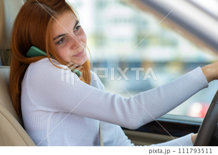 Happy redhead girl talking on her mobile phone behind the wheel driving a car. 111793511