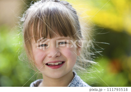 Cute pretty child girl with gray eyes and fair hair smiling in camera outdoors on blurred sunny green and yellow bright bokeh background. 111793518