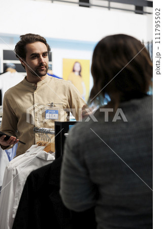 Shopping center employee helping customer and checking apparel in stock using digital tablet. Clothing store consultant offering client assistance in choosing casual outfit Shopping center employee helping customer and checking apparel in stock using digital tablet. Clothing store consultant offering client assistance in choosing casual outfit 111795502