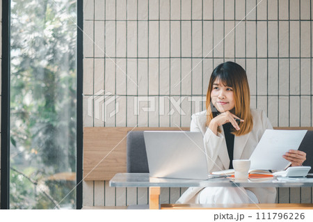 A confident businesswoman assesses documents with a pen in hand, laptop open, in the bright ambiance of a cafe setting. A confident businesswoman assesses documents with a pen in hand, laptop open, in the bright ambiance of a cafe setting. 111796226