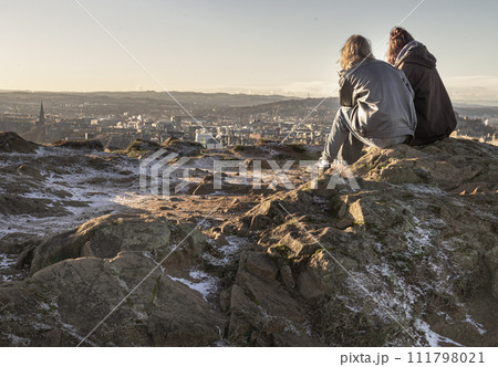 Two young women sitting relaxing on top of rock over the mountain and enjoying looking at scenic view of cityscape. Two young women sitting relaxing on top of rock over the mountain and enjoying looking at scenic view of cityscape. 111798021