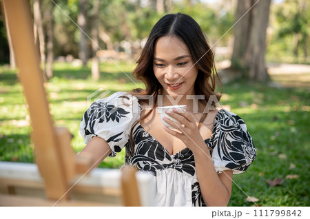 A carefree Asian woman is enjoying hot tea while painting on a canvas on an easel in a park. 111799842