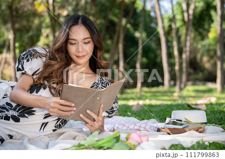 A gorgeous Asian woman is reading a book on a picnic mat while picnicking in a park on a bright day. 111799863