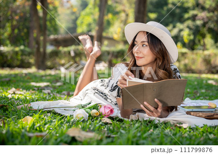 A beautiful and positive Asian woman is lying on a picnic mat and reading a book in a green park. 111799876