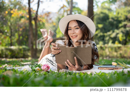 A beautiful and positive Asian woman is lying on a picnic mat and reading a book in a green park. A beautiful and positive Asian woman is lying on a picnic mat and reading a book in a green park. 111799878