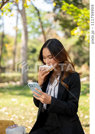 A busy businesswoman is multitasking, eating sandwich in a park and checking emails on her phone. 111799909