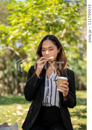 A businesswoman is having a quick breakfast in a city park, eating a sandwich and drinking coffee. A businesswoman is having a quick breakfast in a city park, eating a sandwich and drinking coffee. 111799919
