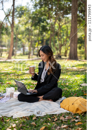 A businesswoman is sipping coffee and working on her digital tablet on a picnic mat in a park. 111799943