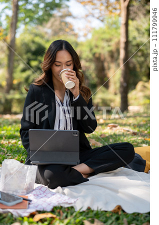 A businesswoman is sipping coffee and working on her digital tablet on a picnic mat in a park. A businesswoman is sipping coffee and working on her digital tablet on a picnic mat in a park. 111799946