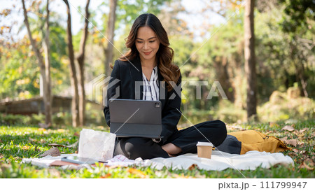 A positive Asian businesswoman remote working at a park, working on her tablet on a picnic mat. A positive Asian businesswoman remote working at a park, working on her tablet on a picnic mat. 111799947