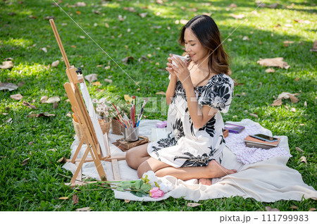 Positive Asian woman is enjoying hot tea and painting on a canvas easel while picnicking in a park. 111799983