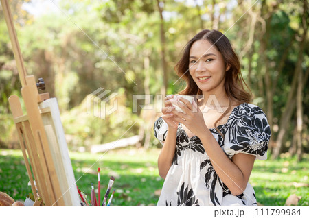 Positive Asian woman is enjoying hot tea and painting on a canvas easel while picnicking in a park. 111799984