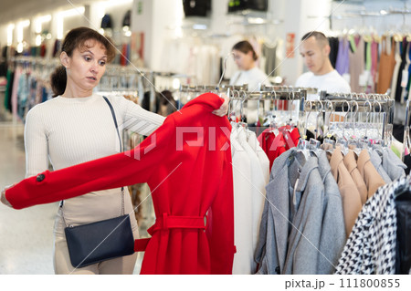 Middle-aged woman choosing unicolorous coat in clothing shop Middle-aged woman choosing unicolorous coat in clothing shop 111800855