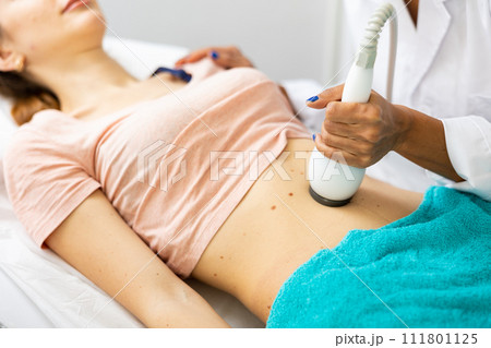 Beautician doing ultrasound lifting procedure in woman's abdominal region 111801125