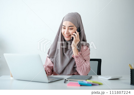 young pretty modern muslim woman in hijab working on laptop in office room, education online, remote work freelancer, using smartphone 111807224