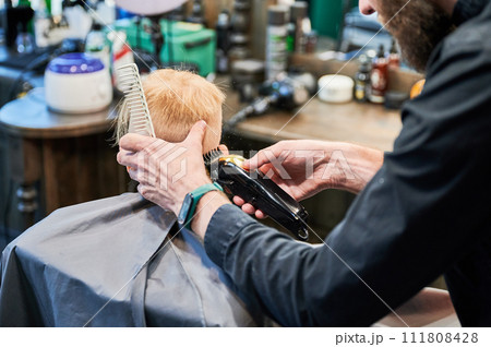 Barber using comb and shaver to cut hair. Professional hairdresser shaving little kid's nape. Barber using comb and shaver to cut hair. Professional hairdresser shaving little kid's nape. 111808428