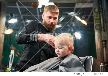 Cute little boy getting haircut. Blond curious kid first time at barbershop. Professional hairdresser making stylish haircut by shaver. Cute little boy getting haircut. Blond curious kid first time at barbershop. Professional hairdresser making stylish haircut by shaver. 111808430
