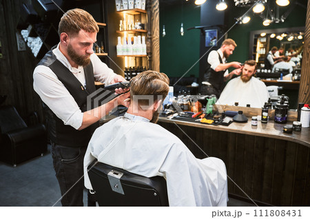 Professional barber using comb and electric shaver to cut side hair. Close-up view of male customer getting stylish haircut. 111808431