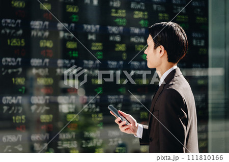 掲示板に映るリアルタイム株価ボードを見てスマホでNISAの取引をする30代のスーツ姿の日本人男性 111810166