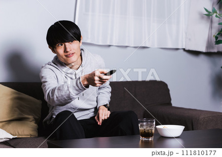 自宅の部屋で深夜にお酒とツマミを肴にテレビドラマやアニメを楽しむ為にリモコンを操作する30代の男性 111810177