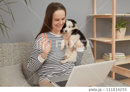 Cheerful friendly Caucasian woman wearing striped shirt sitting on sofa with her puppy dog using laptop computer having video call waving hand showing hello gesture boasting her new pet 111814255