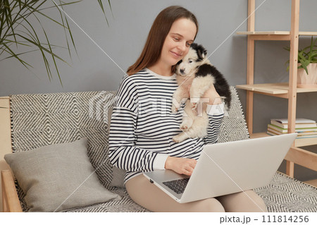Smiling delighted adorable Caucasian woman wearing striped shirt sitting on sofa with her puppy dog using laptop computer typing keyboard looking at notebook display 111814256