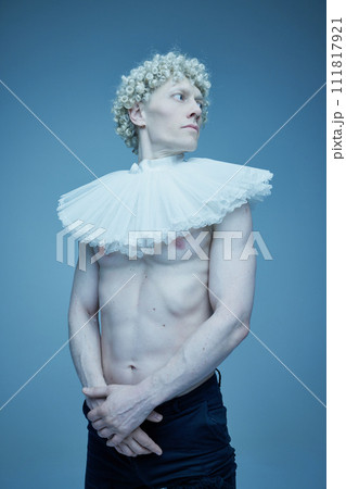 Fashion evolution. Portrait of shirtless man with ruffled collar and black trousers looking away against blue studio background. Fashion evolution. Portrait of shirtless man with ruffled collar and black trousers looking away against blue studio background. 111817921