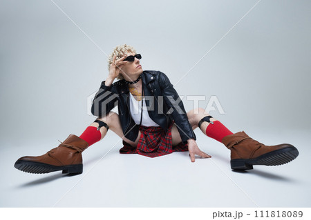 Young man with sunglasses dressed traditional Scottish attire and modern leather jacket posing sitting against grey studio background. Young man with sunglasses dressed traditional Scottish attire and modern leather jacket posing sitting against grey studio background. 111818089