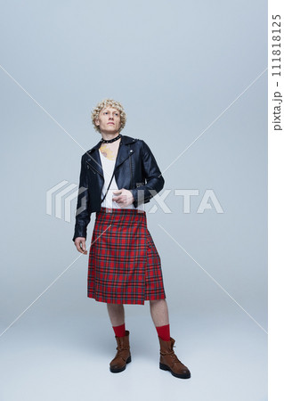 Full-length portrait of young handsome, curly haired man posing in Scottish kilt and leather jacket against grey studio background. 111818125