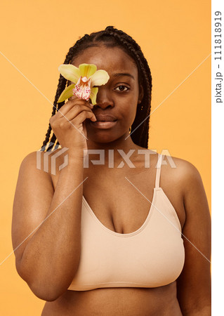 Vertical waist up studio shot of young Black woman wearing bralette covering eye with fresh orchid flower looking at camera Vertical waist up studio shot of young Black woman wearing bralette covering eye with fresh orchid flower looking at camera 111818919