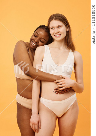 Vertical medium long studio portrait of cheerful gen Z Black woman wearing comfortable lingerie hugging her Caucasian girlfriend 111818950