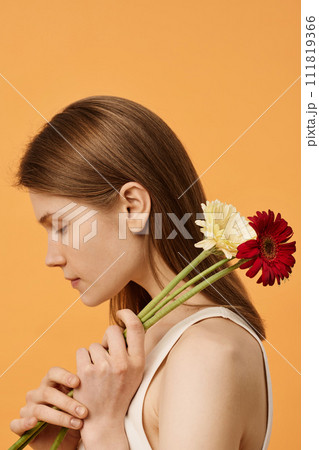 Vertical side view medium close-up studio shot of young Caucasian woman with no makeup posing for camera with fresh gerbera flowers Vertical side view medium close-up studio shot of young Caucasian woman with no makeup posing for camera with fresh gerbera flowers 111819366