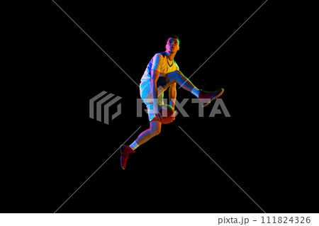 Dynamic portrait of young basketball player dribbling in mid-air against black studio background in mixed neon light. 111824326