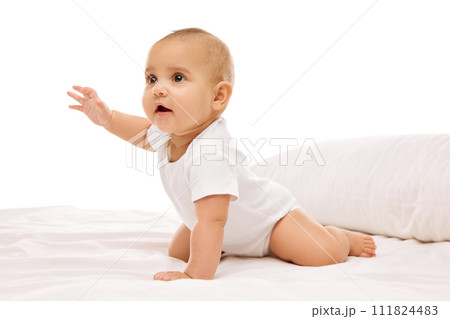 Curious, active, playful little baby girl, child sitting on bed with pillows, making moves, attentively looking against white background Curious, active, playful little baby girl, child sitting on bed with pillows, making moves, attentively looking against white background 111824483