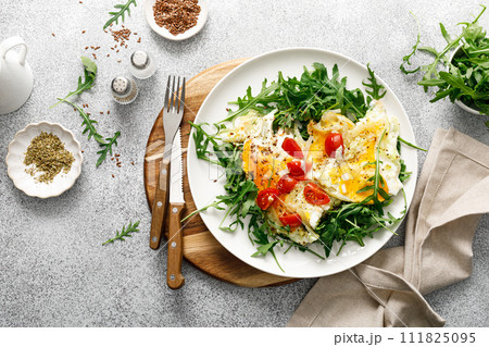 Fried eggs, omelette with onions, tomatoes and fresh arugula salad, top view Fried eggs, omelette with onions, tomatoes and fresh arugula salad, top view 111825095