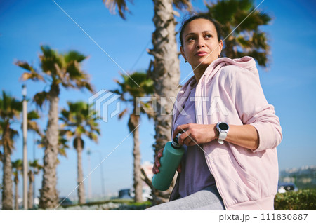 Determined young female athlete, sportswoman holding a steel bottle with refreshing water, relaxing after run outdoor Determined young female athlete, sportswoman holding a steel bottle with refreshing water, relaxing after run outdoor 111830887