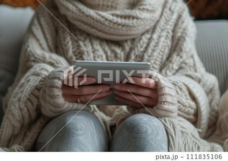 A person cozied up in a knitted sweater and scarf, holding a digital tablet with both hands, the embodiment of comfort and connectivity in a relaxed indoor setting. A person cozied up in a knitted sweater and scarf, holding a digital tablet with both hands, the embodiment of comfort and connectivity in a relaxed indoor setting. 111835106