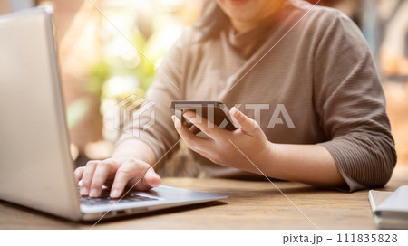 A woman multitasking, using her smartphone and laptop computer, working remotely at a cafe. 111835828