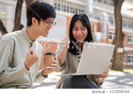 Two cheerful Asian students are celebrating their test scores together while sitting in the park. 111836102