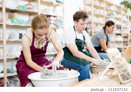 Young woman teacher helps teenagers boy and girl to make product on potter's wheel Young woman teacher helps teenagers boy and girl to make product on potter's wheel 111837132