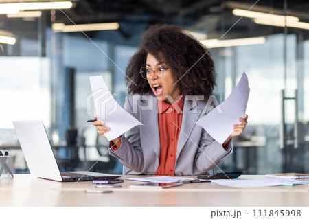 Outraged female with afro hairstyle screaming aggressively while holding documents in hands by desktop with pc. Angry administrator horrified by workload with paperwork at beginning of busy day. Outraged female with afro hairstyle screaming aggressively while holding documents in hands by desktop with pc. Angry administrator horrified by workload with paperwork at beginning of busy day. 111845998