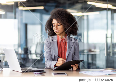 Focused female in round eyeglasses looking at pc screen while filling in document on writing board in office interior. Confident entrepreneur copying information from email to printed papers. 111846026