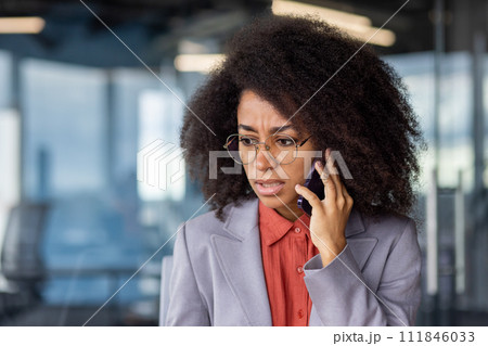Worried african american lady with bushy hairstyle holding smartphone by ear and listening bad news. Disturbed office worker having stressful distant conversation by mobile gadget during daytime. Worried african american lady with bushy hairstyle holding smartphone by ear and listening bad news. Disturbed office worker having stressful distant conversation by mobile gadget during daytime. 111846033